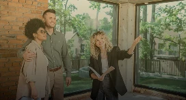 A real estate agent from Lyon Property Group gestures and smiles while showing a house to a couple standing together in an unfinished room with large windows and exposed brick walls in Rhode Island and Eastern Connecticut.