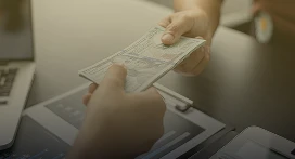 A person hands a stack of U.S. dollar bills to another across a desk, with a laptop and documents in the background—capturing a transaction typical in property management by Lyon Property Group in Rhode Island and Eastern Connecticut.