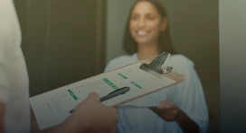 A person holds a clipboard with a form and pen, while a smiling woman in the background stands at a doorway—ready to respond or sign—for Lyon Property Group, your trusted property management team in Rhode Island and Eastern Connecticut.