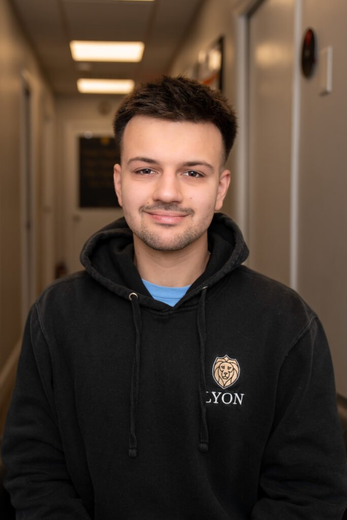 A young man with short dark hair sits indoors, smiling softly. He wears a black hoodie with a lion emblem and the words "Lyon Property Group." The background shows a hallway with beige walls and ceiling lights in Rhode Island and Eastern Connecticut.