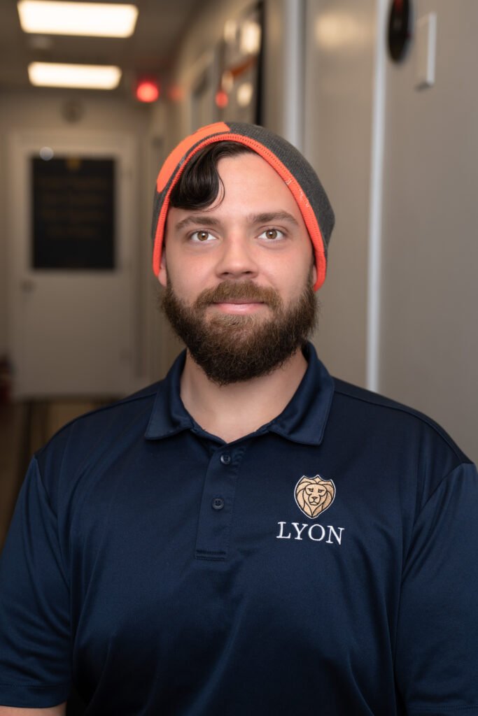 A man with a beard and mustache, wearing a gray and orange beanie and a navy blue Lyon Property Group polo shirt, sits indoors in a brightly lit hallway, representing property management in Rhode Island and Eastern Connecticut.