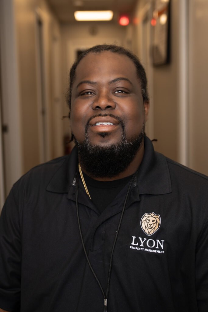 A man with a beard and mustache smiles at the camera while wearing a black Lyon Property Management polo shirt, standing in a well-lit hallway with white walls and doors, representing Rhode Island and Eastern Connecticut.