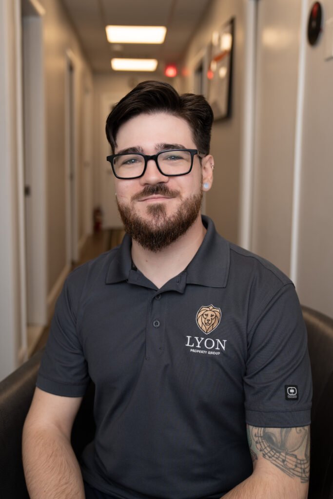 A man with dark hair, glasses, and a beard sits in a hallway, smiling. He wears a gray Lyon Property Group polo shirt, representing property management in Rhode Island and Eastern Connecticut. A tattoo shows on his left forearm.