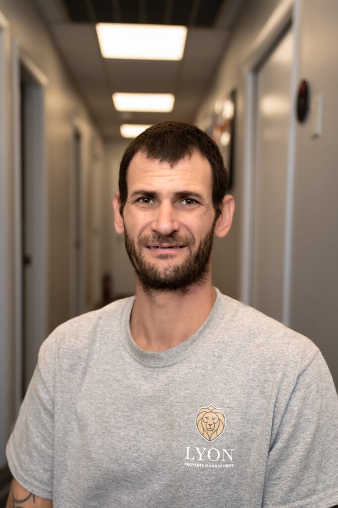 A man with short dark hair and a beard, wearing a gray T-shirt with a Lyon Property Management logo, sits and smiles in a brightly lit hallway—proudly representing Rhode Island and Eastern Connecticut.