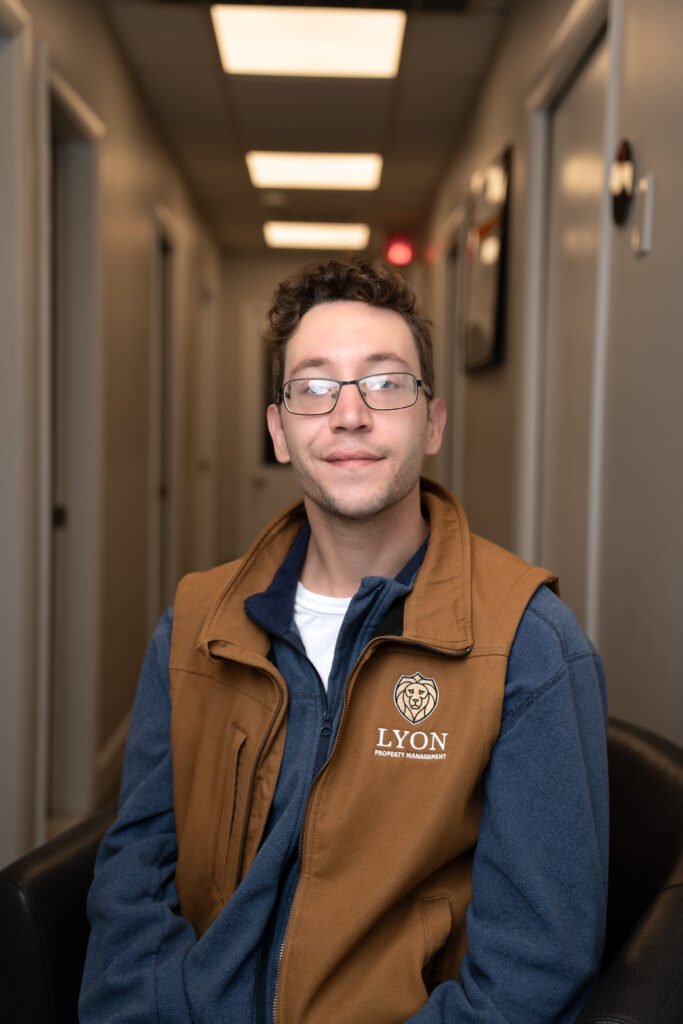 A person with short brown hair and glasses sits in a hallway, wearing a brown and blue vest with a Lyon Property Group logo. The well-lit hallway features closed doors, representing property management professionalism in Rhode Island and Eastern Connecticut.