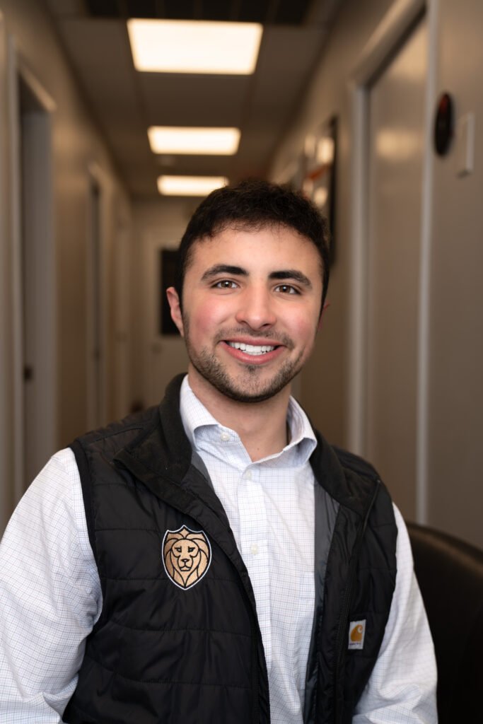 A young man with short dark hair and a beard smiles while sitting indoors. He wears a white button-up shirt and a black Lyon Property Group vest, representing expert property management in Rhode Island and Eastern Connecticut. The background shows a hallway with bright lights.