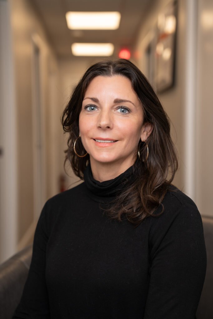 A woman with wavy brown hair, wearing a black turtleneck and gold hoop earrings, sits and smiles in a well-lit hallway with framed pictures, reflecting the welcoming atmosphere of Lyon Property Group’s Property Management team.