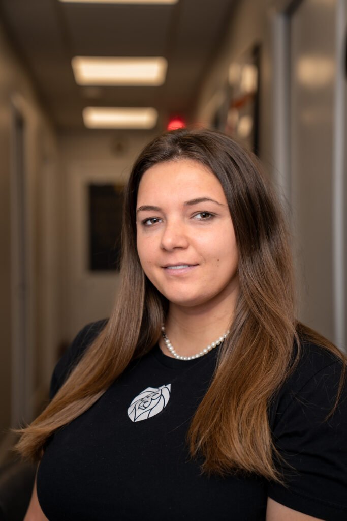 A woman with long brown hair wearing a black shirt with a rose graphic and pearl necklace stands in a well-lit hallway, reflecting the professionalism of Lyon Property Group, leaders in Rhode Island and Eastern Connecticut Property Management.