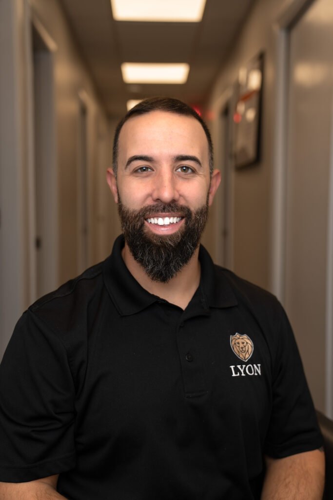A man with a dark beard and short hair, wearing a black polo shirt with a bear logo and the words Lyon Property Group, smiles while sitting in a brightly lit hallway—proudly representing property management in Rhode Island and Eastern Connecticut.