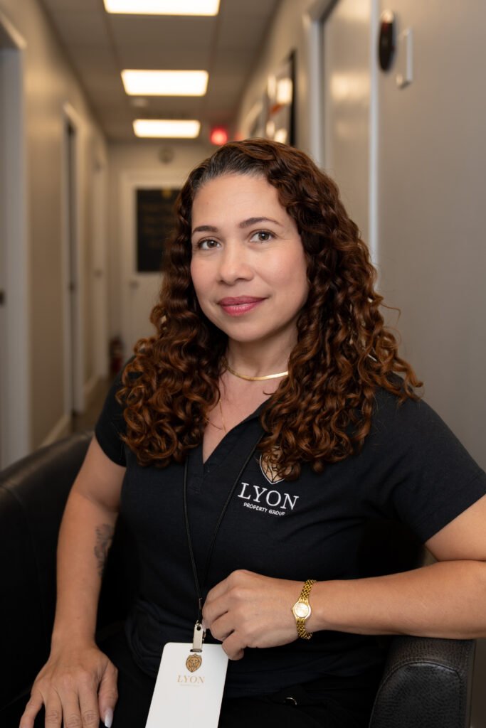 A woman with curly hair sits in a hallway, wearing a black Lyon Property Group polo shirt and gold jewelry, holding a badge. The setting reflects the professional property management services offered across Rhode Island and Eastern Connecticut.