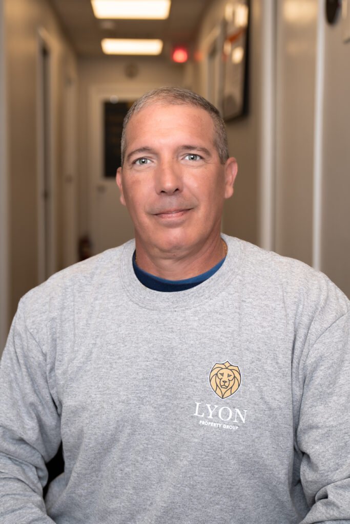 A man with short gray hair sits indoors, wearing a light gray Lyon Property Group sweatshirt. The setting, ideal for Property Management in Rhode Island and Eastern Connecticut, features a hallway with white walls and framed pictures.