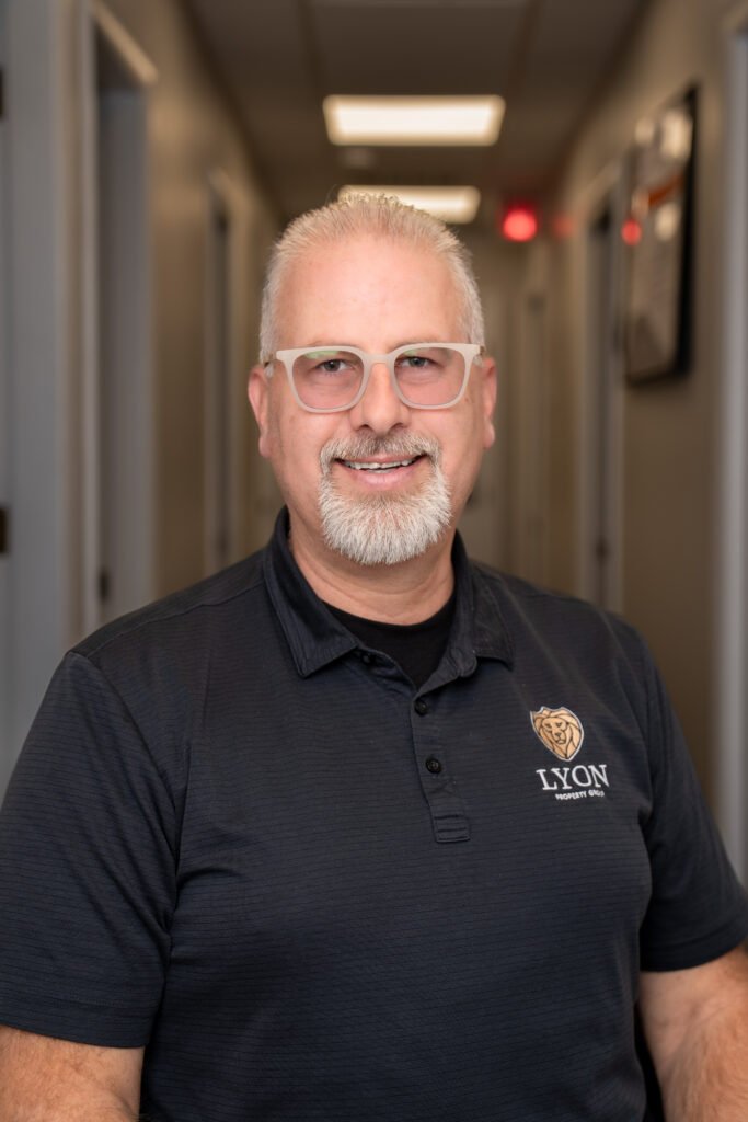 A middle-aged man with short gray hair, a goatee, and clear glasses smiles while standing in a hallway. He is wearing a black polo shirt with the Lyon Security Inc. logo, part of Lyon Property Group serving Rhode Island and Eastern Connecticut.