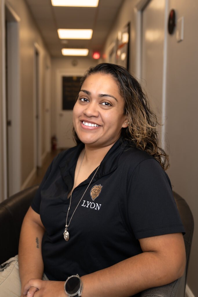 A smiling woman with wavy hair sits in a hallway on a dark chair, wearing a black Lyon Property Group polo shirt, light pants, and a pendant necklace. The hallway features bright ceiling lights and light-colored walls.