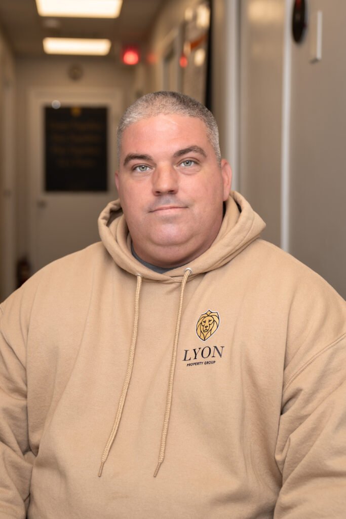 A man with short gray hair wearing a beige Lyon Property Group hoodie sits in a softly lit hallway, looking at the camera. The scene hints at professional property management in Rhode Island and Eastern Connecticut.