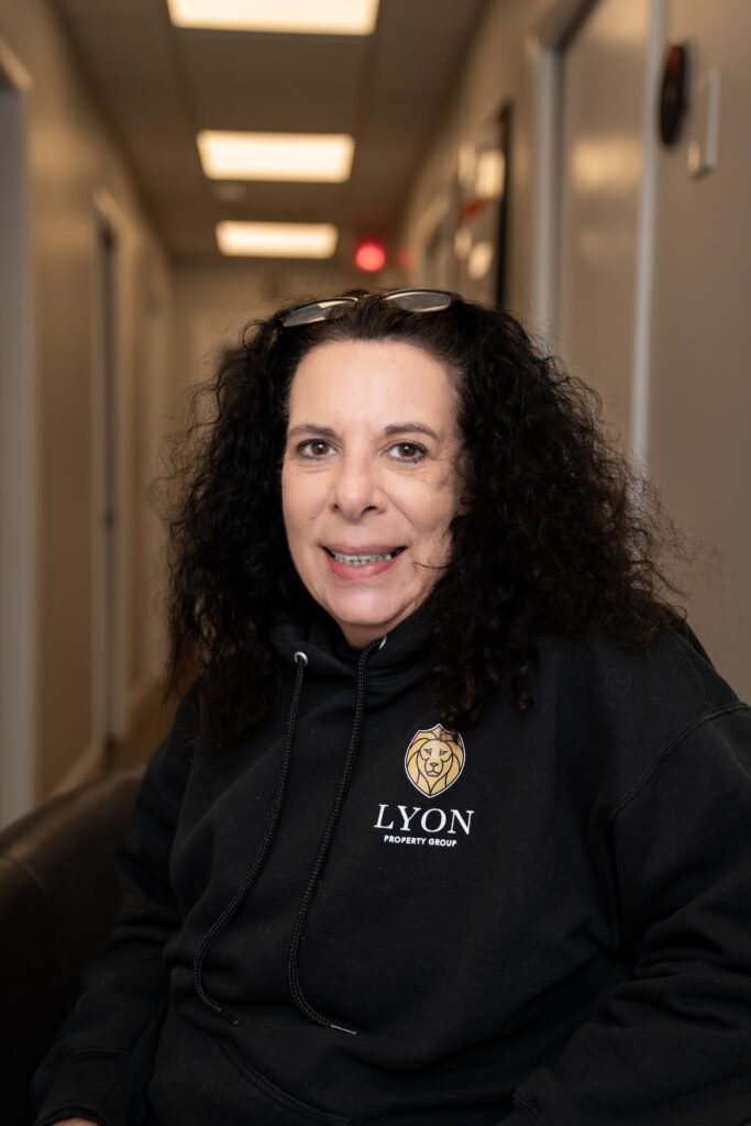 A smiling person with curly dark hair sits indoors, wearing a black Lyon Property Group hoodie—representing expert property management in Rhode Island and Eastern Connecticut—with glasses resting on their head. The background shows a hallway with overhead lights.