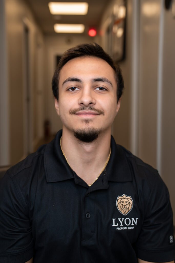 A young man with short dark hair and a trimmed beard, wearing a black Lyon Property Group polo, is seated in a softly lit hallway with neutral-colored walls—representing professional property management across Rhode Island and Eastern Connecticut.