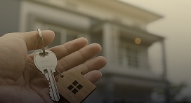 A hand holding house keys with a wooden keychain shaped like a house; blurred background shows a modern home. Lyon Property Group specializes in property management across Rhode Island and Eastern Connecticut.