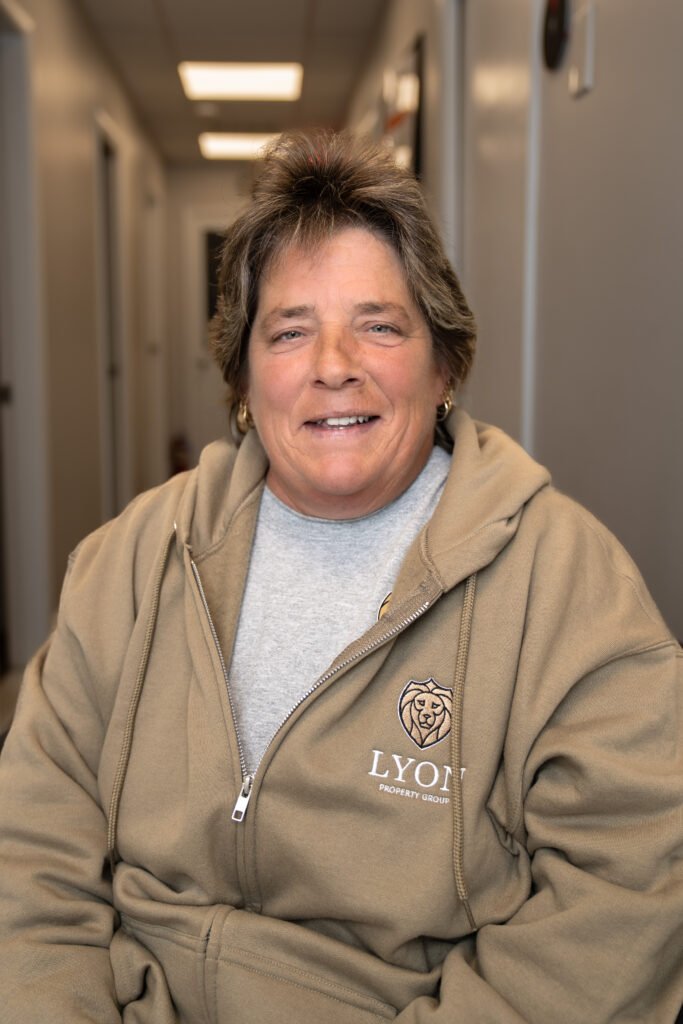 A person with short brown hair is smiling and wearing a tan zip-up hoodie with the Lyon Property Group logo, representing Property Management in Rhode Island and Eastern Connecticut, sitting in a hallway with doors and ceiling lights visible.