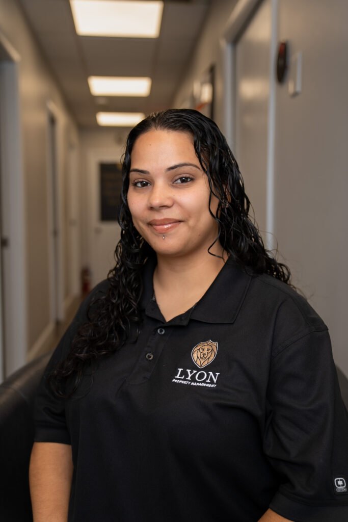 A woman with long dark hair and a lip piercing smiles while wearing a black Lyon Property Group polo shirt, standing in a brightly lit hallway with white walls and ceiling lights in Rhode Island and Eastern Connecticut.
