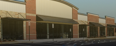 A row of empty retail storefronts with large glass windows and brick exteriors in a shopping plaza, managed by Lyon Property Group, serving Rhode Island and Eastern Connecticut, with a mostly empty parking lot in front.