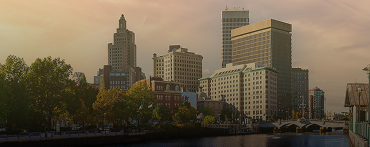 A city skyline at sunset with tall office buildings, autumn trees, and a river in the foreground—capturing the landscape Lyon Property Group manages across Rhode Island and Eastern Connecticut. The sky is softly lit with warm colors.