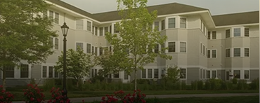Three-story beige apartment building with white trim and many windows, surrounded by trees, bushes, and flowers, with a black lamp post in front—professionally managed by Lyon Property Group in Rhode Island and Eastern Connecticut.