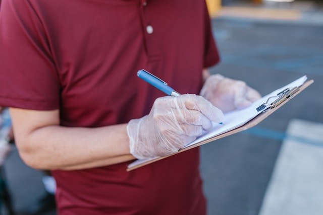 A person in a red shirt and clear disposable gloves writes on a clipboard with a blue pen, standing outdoors—representing Lyon Property Group’s commitment to quality property management in Rhode Island and Eastern Connecticut.